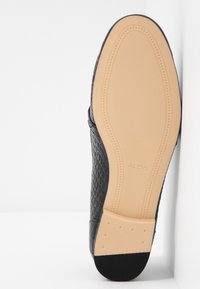 Chaussure plate noire texturée avec une semelle en caoutchouc, dotée d'une semelle intérieure lisse de couleur claire et d'un marquage subtil embossé sur la partie inférieure.