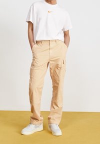 Beige cargobyxor med flera fickor, som bärs med en vit t-shirt med en orange Nike-logga och vita sneakers. Slät tygkvalitet.
