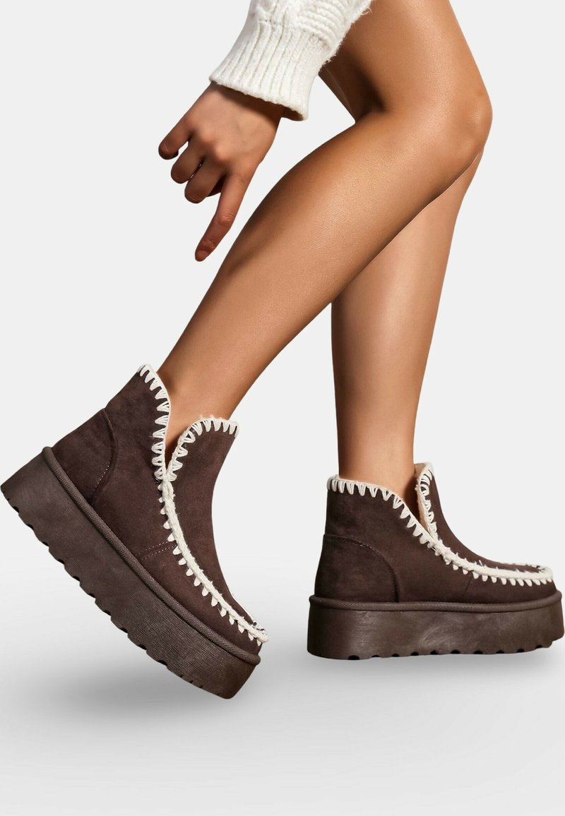 Des jambes portant des bottines à plateforme en daim marron foncé avec des accents de couture blanche, une semelle surélevée et une manche de pull blanche visible sur la main.