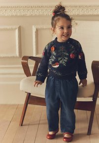 Strickpullover in Marineblau mit floralem Stickmuster, kombiniert mit lockeren blauen Denim-Hosen und schimmernden roten Schuhen, steht neben einem Holzstuhl.