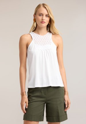 Vrouw die een witte mouwloze top met kanten details draagt en olijfgroene opgerolde shorts, geaccentueerd met gouden armbanden en grote oorbellen.