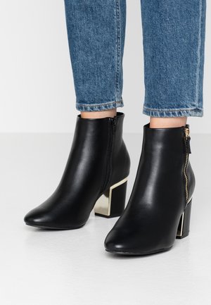 Stivali neri in pelle alla caviglia con punta appuntita, cerniera laterale e tacco quadrato con un accento in oro metallico. Indossati con jeans blu.