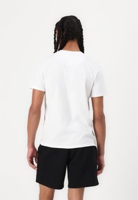 White cotton t-shirt with short sleeves, visible stitching at the neckline, paired with black shorts, viewed from the back.