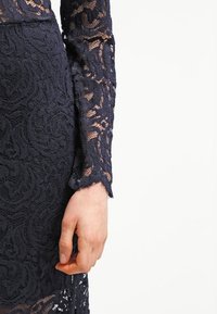 Bras droit et main d'une personne portant une robe en dentelle bleu marine ajustée avec des manches longues transparentes et des motifs floraux.