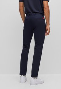 Navy blue slim-fit trousers with a flat waistband and two back pockets, paired with white sneakers for a casual look.