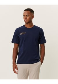 Navy modrý bavlněný tričko s krátkým rukávem a kulatým výstřihem. Na levé hrudi má zlaté logo "Les Deux". Jednoduchý design, hladká textura.