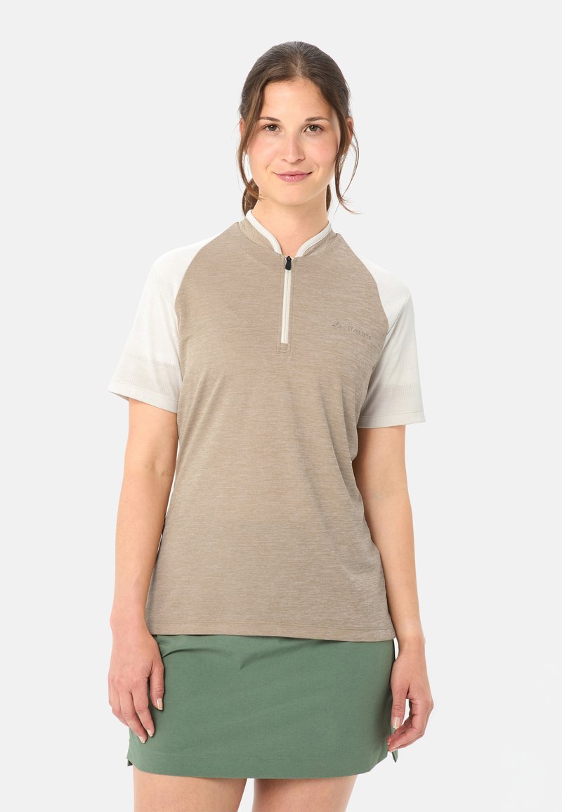 Femme aux cheveux bruns attachés en queue de cheval, portant une chemise beige et blanche à fermeture éclair et une jupe verte, debout devant un fond blanc.