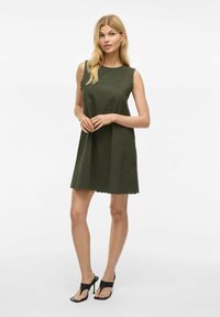 Vestido verde oliva con dobladillo en forma de concha y diseño sin mangas, hecho de tela texturizada. Usado con sandalias de tacón negro.