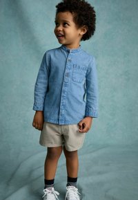 Camisa de mezclilla con botones y bolsillo, combinada con pantalones cortos de khaki claros. Zapatillas blancas con calcetines a rayas, de pie frente a un fondo texturizado.