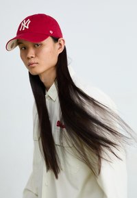 Red baseball cap with white "NY" logo, worn over long dark hair. White button-up shirt with red embroidery. Smooth texture, casual design.