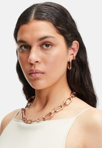 Guldfarvet kædedesign lavet af sammenflettede led, med en glat overflade. Modellen bærer små hoops øreringe i en matchende farve.