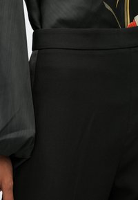 Calças pretas com uma textura suave, apresentando um cós largo e uma silhueta ajustada. Acompanhadas por uma blusa transparente de tom cinza escuro.