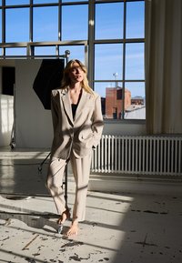 Femme blonde pieds nus en costume beige se tient dans un studio baigné de lumière près d'une grande fenêtre, avec du matériel de photographie et un paysage urbain visibles à l'extérieur.