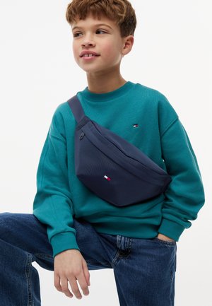 Young boy wearing teal sweatshirt and blue jeans with navy crossbody bag, sitting with one hand in pocket against white background.