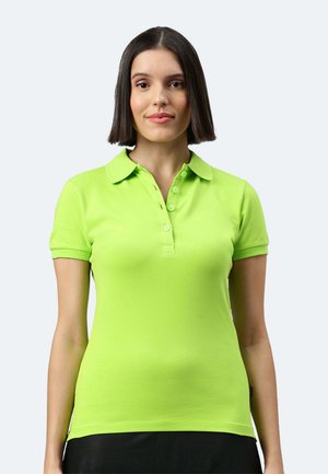 Junge Frau mit glatten dunklen Haaren, die ein hellgrünes Polohemd mit kurzen Ärmeln trägt und vor einem schlichten hellen Hintergrund steht.