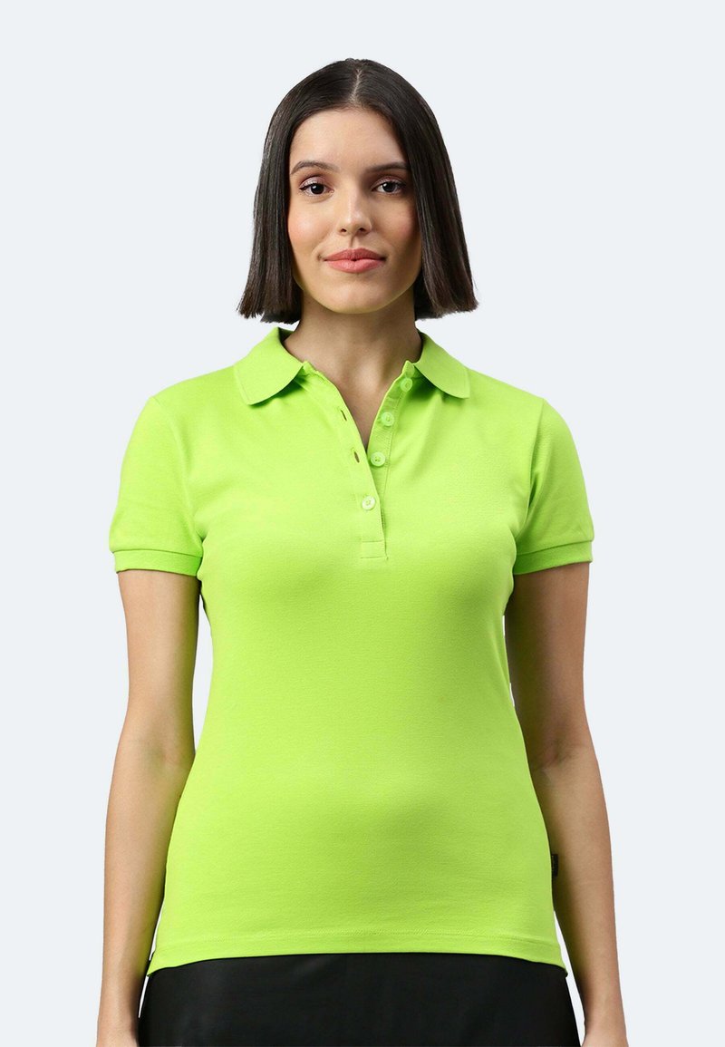 Jeune femme aux cheveux bruns raides portant un polo à manches courtes vert vif, debout devant un fond clair uni.