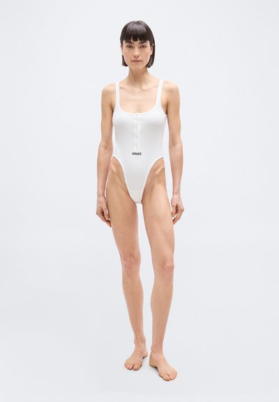 Γυναίκα με κοντά σκούρα μαλλιά που φοράει λευκό κορμάκι με κουμπιά και λογότυπο "Versace" σε απλό φωτεινό φόντο.