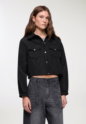 Giacca nera trapuntata, cropped, con colletto, due tasche sul petto, bottoni a pressione argentati e polsini elasticizzati, abbinata a pantaloni grigi a vita alta.