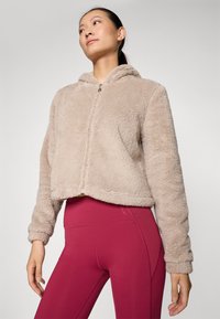 Veste en polaire beige coupée avec une capuche et une fermeture éclair à l'avant, dotée d'un tissu texturé et de poignets élastiques, associée à des leggings roses taille haute.