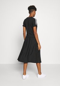 En person står med ryggen mot kameran, iklädd en svart kortärmad skjorta med vita ränder, en svart midiklänning och vita sneakers på en vit bakgrund.