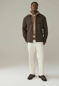 Brun skjorta över en beige hoodie, vita jeans och mörka loafers. Jackan har två bröstfickor och en spetsig krage.