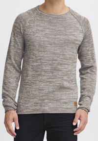 Pull en mailles marbrées brunes avec un col rond, des manches longues, des poignets côtelés et une petite étiquette en cuir au bas. Texture douce.