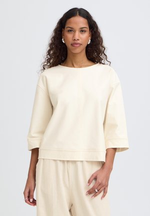 Žena s kudrnatými vlasy v nadměrně velkém krémovém topu s rukávy do loktů a odpovídajících krémových kalhotách stojí před jednoduchým světlým pozadím.