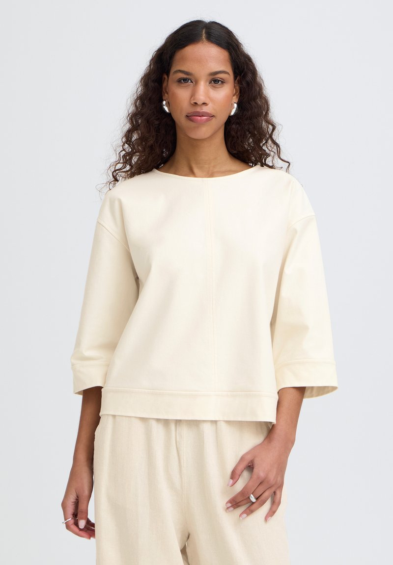 Lokkis juustega naine kannab üle suuruses kreemikat pikka varrukateta pluusi ja sobivaid kreemikaid pükse, ta seisab ühevärvilise heleda tausta ees.