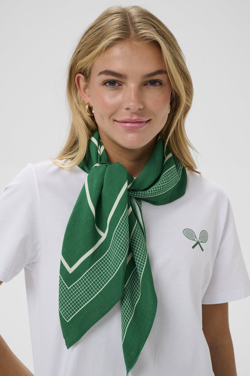 Sciarpa verde con un motivo geometrico, caratterizzata da accenti bianchi. Indossata in modo sciolto attorno al collo, abbinata a una maglietta bianca.