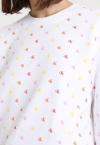 Maglietta bianca a girocollo con un motivo ripetuto di piccoli loghi "cK" in rosso, arancione, giallo e rosa su tutta la superficie.