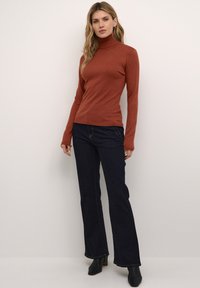 Rostfarbener Rollkragenpullover mit langen Ärmeln, kombiniert mit dunkelblauen, ausgestellten Jeans. Der Stoff wirkt weich und hat eine glatte Textur. Schwarze Stiefeletten mit Absatz.