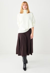 Weißer Rollkragenpullover mit lockerer Passform, kombiniert mit einem dunkelbraunen, plissierten Midirock und schwarzen, kniehohen Stiefeln mit spitzen Zehen.