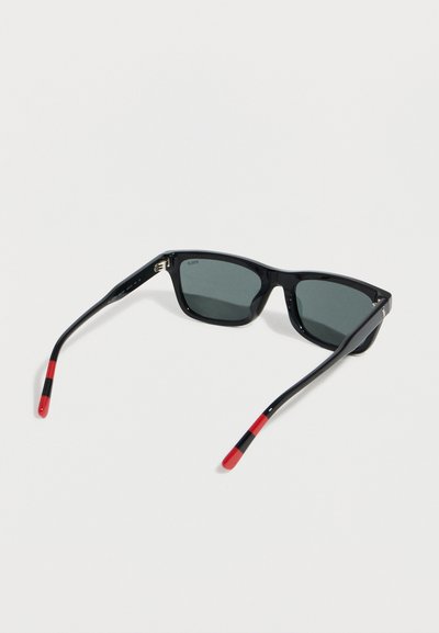 Μαύρα ορθογώνια γυαλιά ηλίου με σκουρόχρωμους φακούς και μαύρους βραχίονες που έχουν κόκκινες άκρες και μια λεπτή μαύρη ρίγα κοντά στα άκρα.