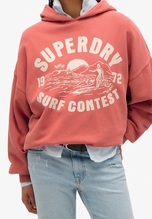 Osoba ubrana w koraloworóżową bluzę z napisem "SUPERDRY SURF CONTEST 1972" i grafiką surfingową, w połączeniu z jasnoniebieskimi dżinsami i koszulą pod spodem.