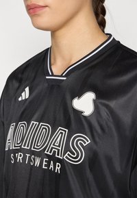 Detailní záběr na osobu v černém sportovním drese Adidas s bílými logy a pruhovaným límcem, mírně otočenou vlevo.