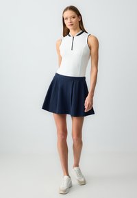 Vrouw staande in een mouwloze witte rits-top, marineblauwe geplooide rok en witte sneakers tegen een egale lichte achtergrond.