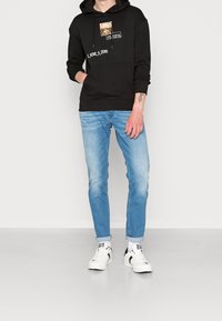 Czarna bluza z kapturem z grafiką z przodu, zestawiona z jasnoniebieskimi obcisłymi dżinsami i białymi tenisówkami z czarnymi detalami.