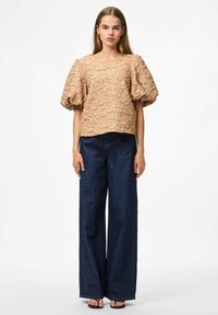 Top beige con maniche a palloncino e motivo floreale testurizzato, abbinato a pantaloni a gamba larga in denim scuro e sandali neri con tacco.