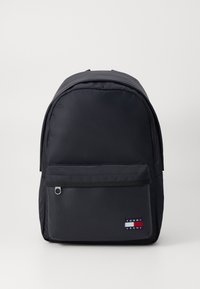 Sac à dos en nylon noir avec une forme arrondie, une poche zippée à l'avant et un petit logo en patch aux couleurs rouge, blanc et bleu. Texture lisse.
