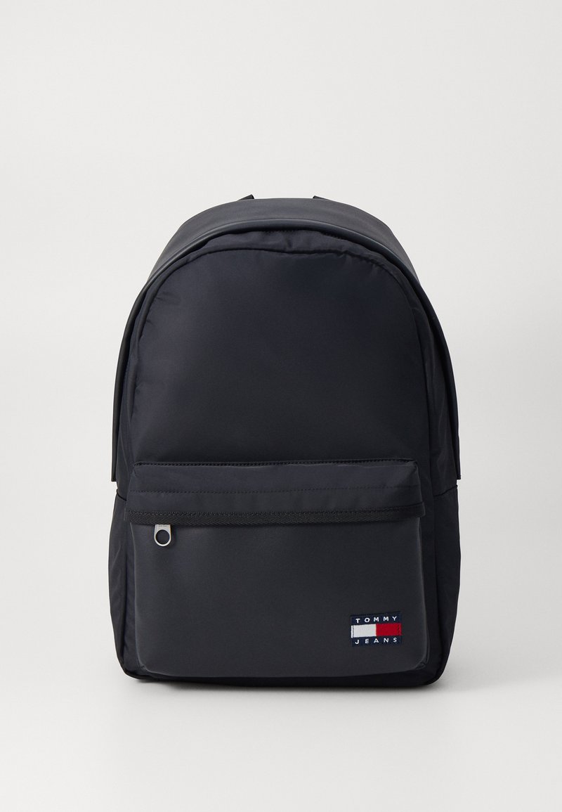 Sac à dos en nylon noir avec une forme arrondie, une poche zippée à l'avant et un petit logo en patch aux couleurs rouge, blanc et bleu. Texture lisse.