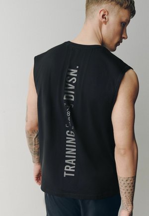 Ung mand med tatoveringer iført en sort ærmeløs skjorte med lodret "TRAINING DIVSN." tekst på ryggen, vendt væk fra kameraet.