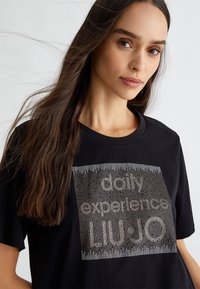 T-shirt in cotone nero con maniche corte, che presenta una grafica testuale metallica argentata con la scritta "esperienze quotidiane LIU-JO." Texture liscia, vestibilità rilassata.