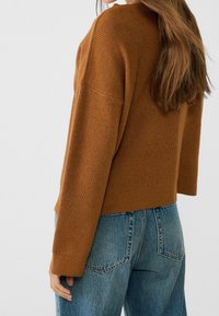 Pull marron tricoté à coupe décontractée, épaules tombantes et poignets côtelés, associé à un jean bleu clair taille haute.