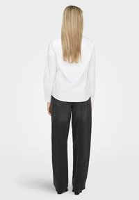 Femme blonde debout, dos à la caméra, portant une chemise blanche à manches longues et un jean noir ample, sur un fond uni.