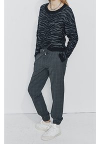 Pull en noir et gris à rayures zébrées avec un col rond, associé à un jogging à carreaux gris, avec une taille élastique et des poignets, porté avec des baskets blanches.