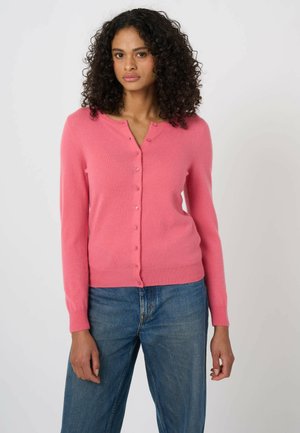 Vrouw met krullend haar, gekleed in een roze vest met knopen en een blauwe spijkerbroek, staand voor een effen lichte achtergrond.