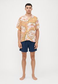Camisa de manga corta con botones y un patrón de batik en naranja y verde, combinada con pantalones cortos azul marino. Tela ligera, ajuste holgado.