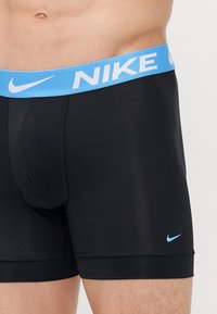 Čierne boxerské trenky s svetlomodrým elastickým pásom, na ktorom sa nachádza biele logo Nike. Tkanina vyzerá hladko a pružne.