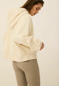 Kremowa bluza z kapturem, o luźnym kroju i ściągaczami na rękawach, zestawiona z teksturowanymi, brązowymi legginsami. Widoczny miękki, casualowy materiał.