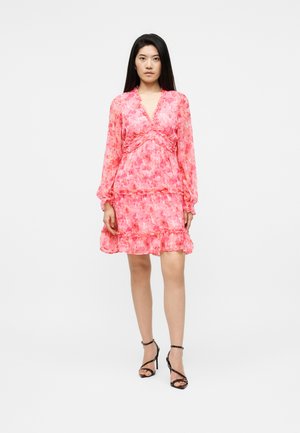 Femme portant une robe rose à fleurs à manches longues avec des volants, debout, associée à des sandales à talons hauts noires à brides sur un fond uni.